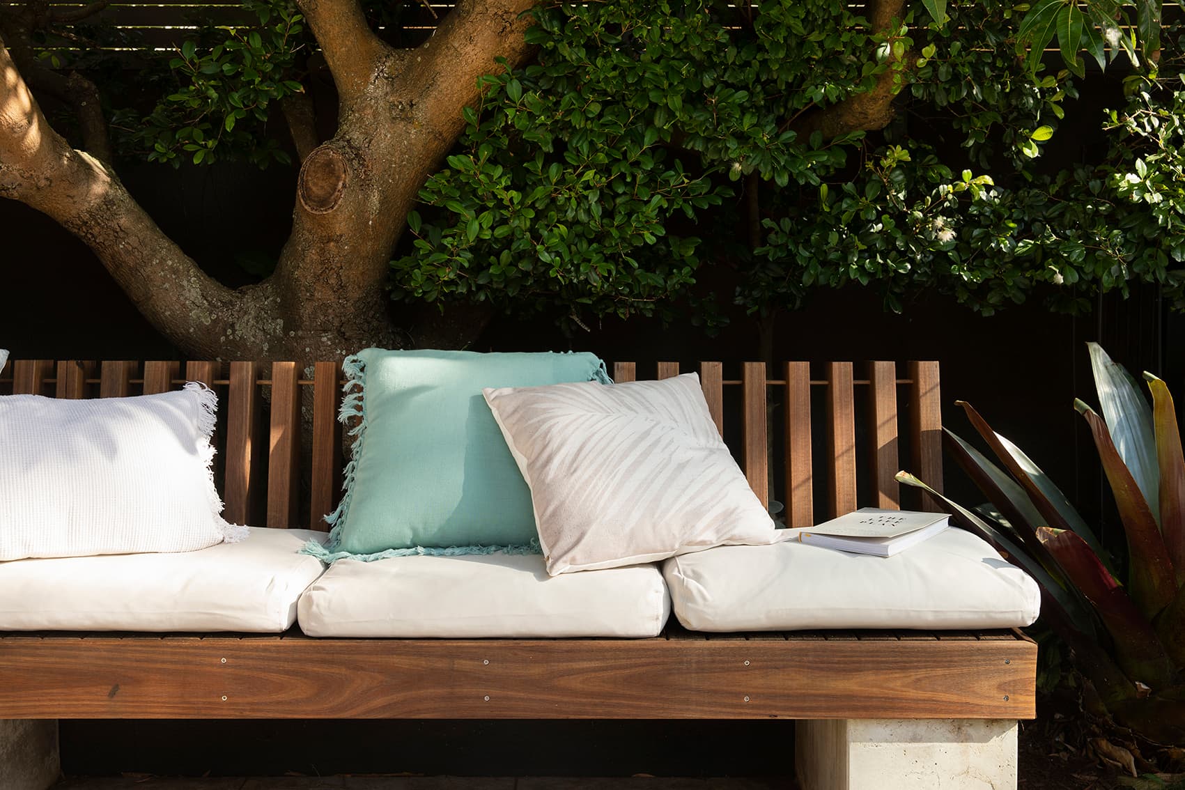 Timber slat bench with white and pale green cushions beneath a spreading tree