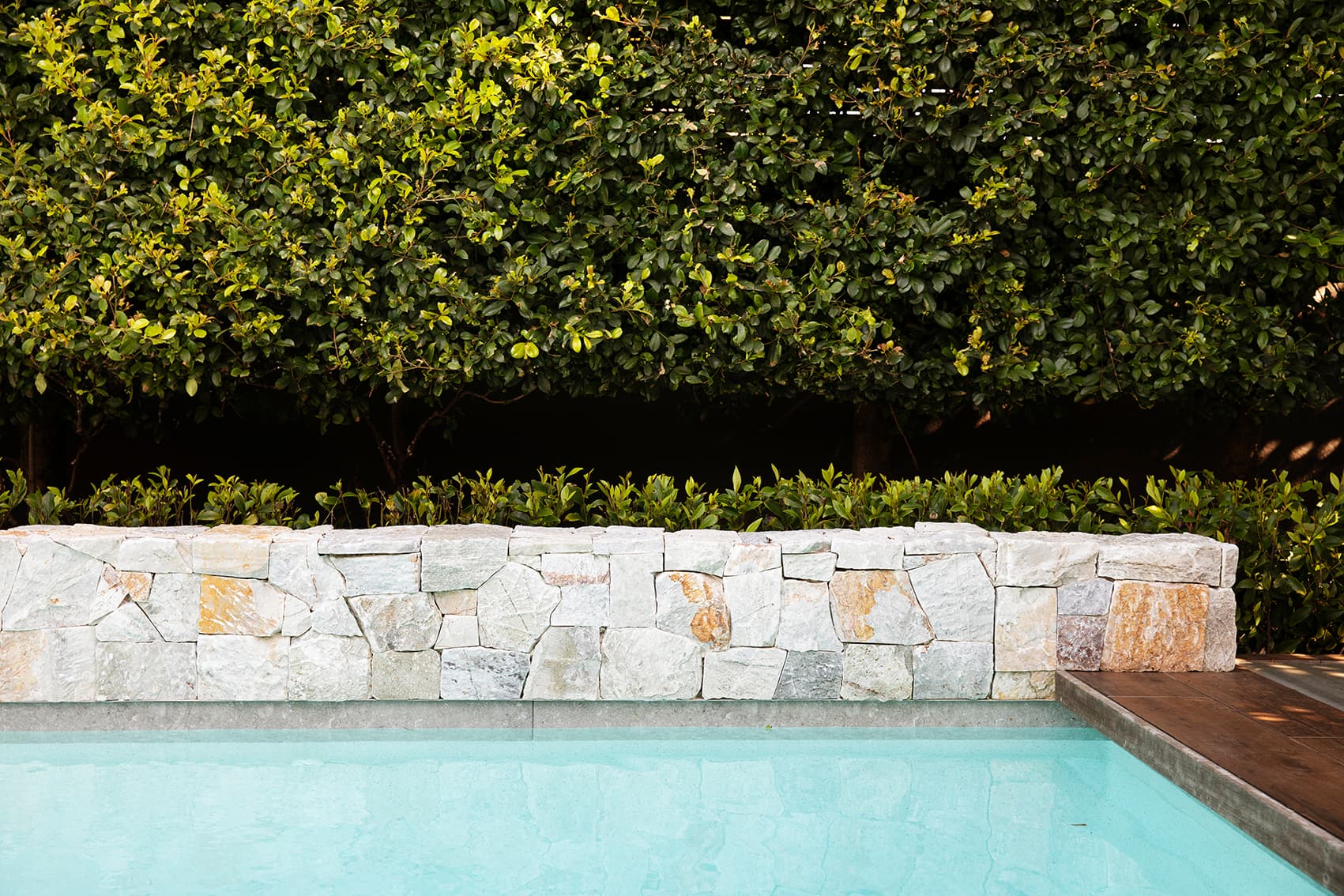 Sandstone clad pool wall backed by a clipped green hedge above still blue water