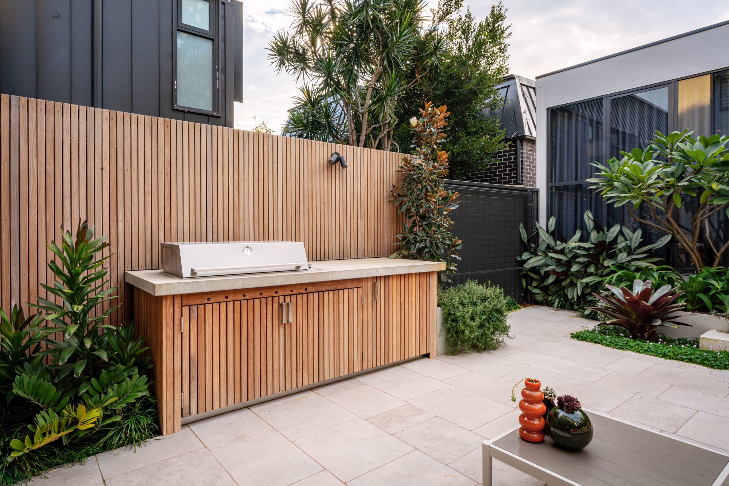 Blackbutt outdoor kitchen with a concrete benchtop beside limestone paving and garden planting.