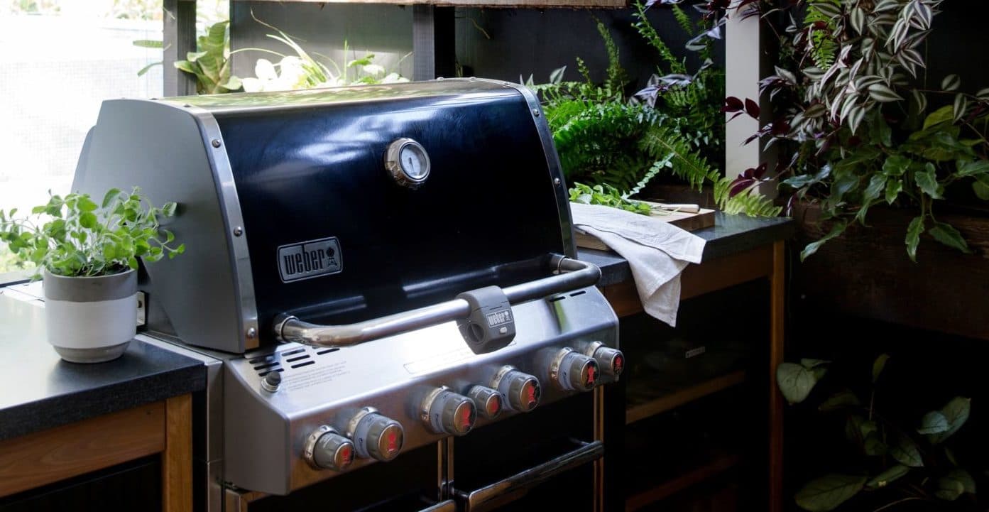 Stainless steel Weber barbecue built into a stone-topped outdoor kitchen among leafy greenery