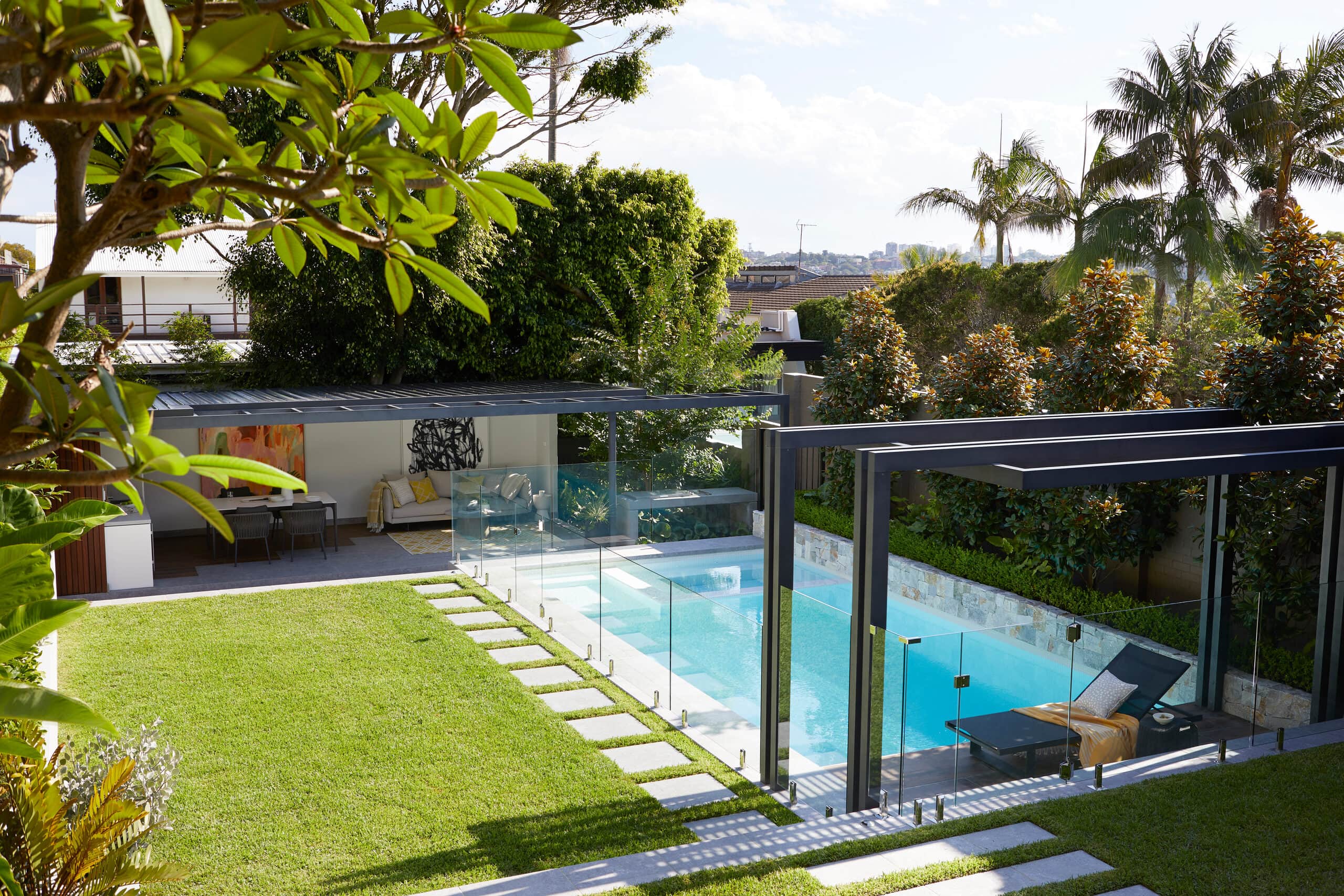 Elevated view over a tropical backyard pool with glass fencing lawn and an open pavilion framed by palms