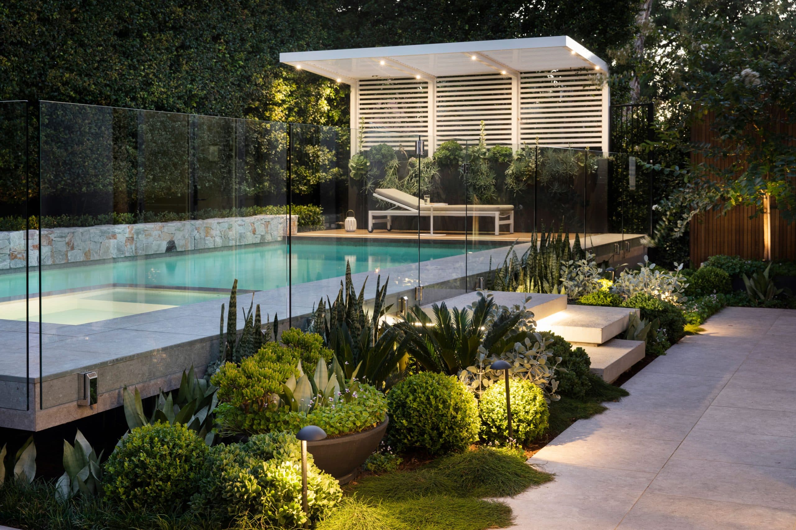 Pool and pavilion glowing at dusk with uplit planting and floating steps along the path