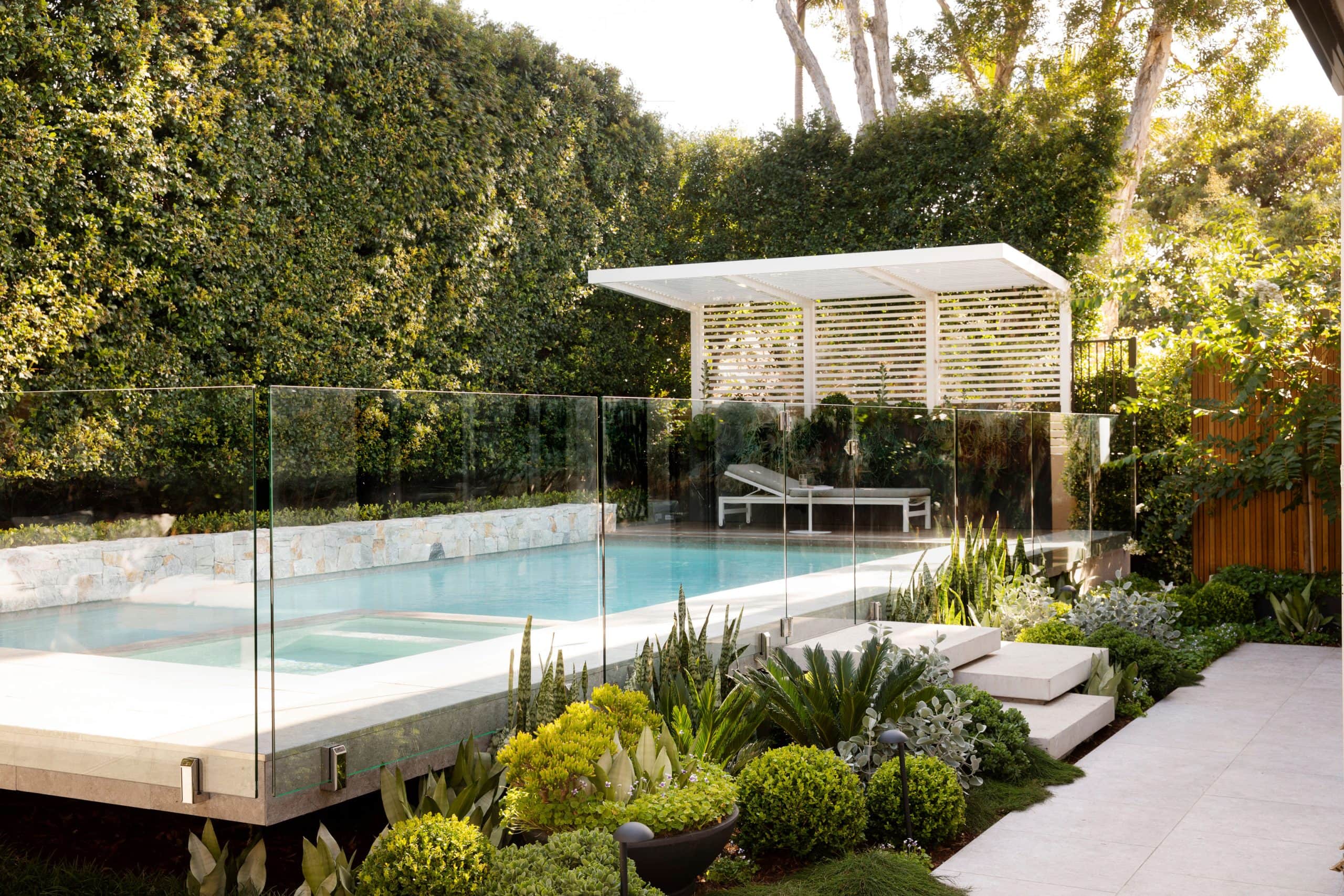 Floating pool and spa behind glass fencing with white slatted pergola and layered native garden at dusk